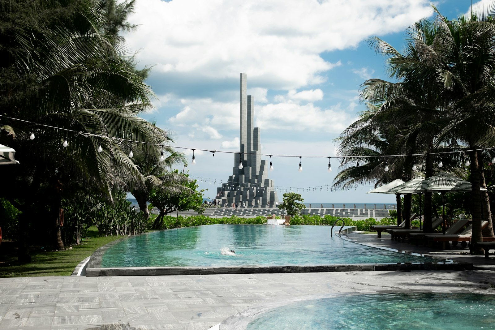 Resort infinity pool framed by palm trees overlooking the ocean in Da Nang, with the APEC Park monument beyond